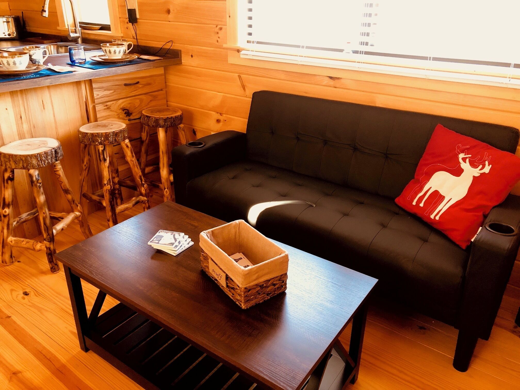 A cozy cabin interior with a wooden dining table, a black sofa with a red pillow, and a small kitchen area.