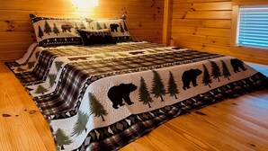 Ein gemütliches Schlafzimmer in einer Holzhütte mit einem Bett, das eine Bettdecke mit Waldmotiv, Holzwänden und einem Fenster mit Jalousien zeigt.