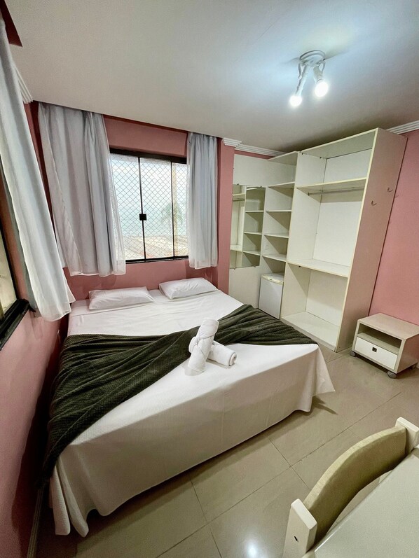 A neatly made bed with a green blanket, white curtains, and a towel on the side. There is a white wardrobe with shelves and a small bedside table. - Webi Coliving - Coqueiros (Florianópolis)