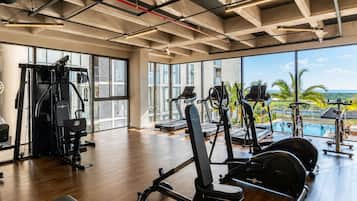 Un gimnasio con equipo de ejercicio, ventanas amplias y vistas a una piscina y palmeras.
