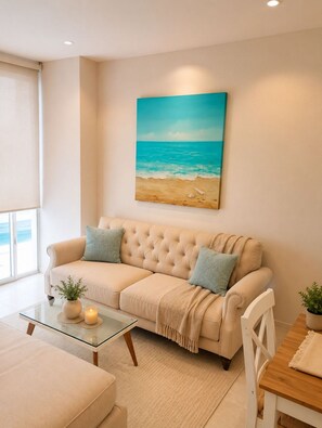 Een woonkamer met een beige bank, een glazen salontafel, een houten eettafel, en een schilderij van een strand.