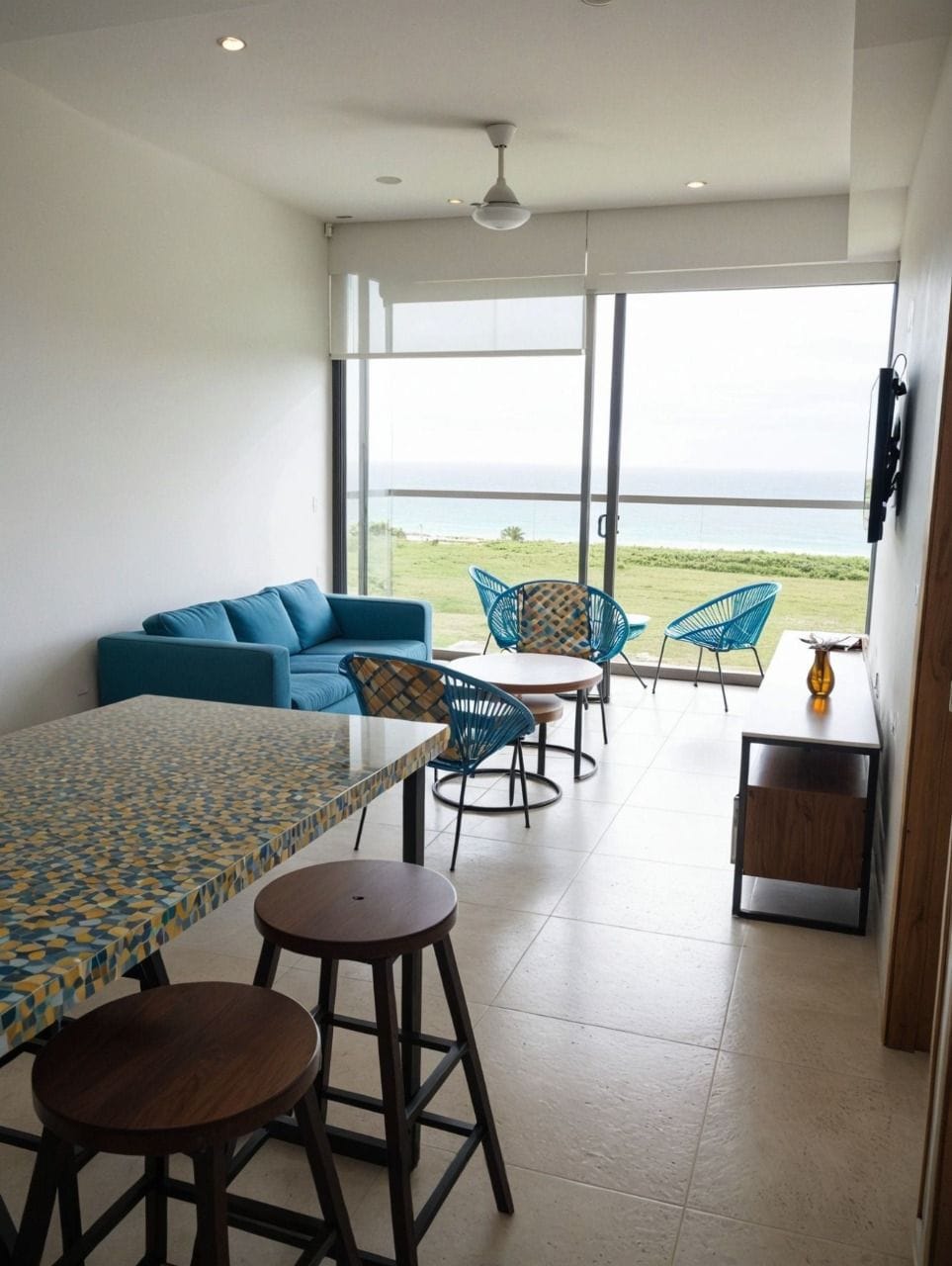 A modern living room with a sofa, a dining table with stools, and two chairs. A TV is mounted on the wall, and there is a view of the ocean through large windows.