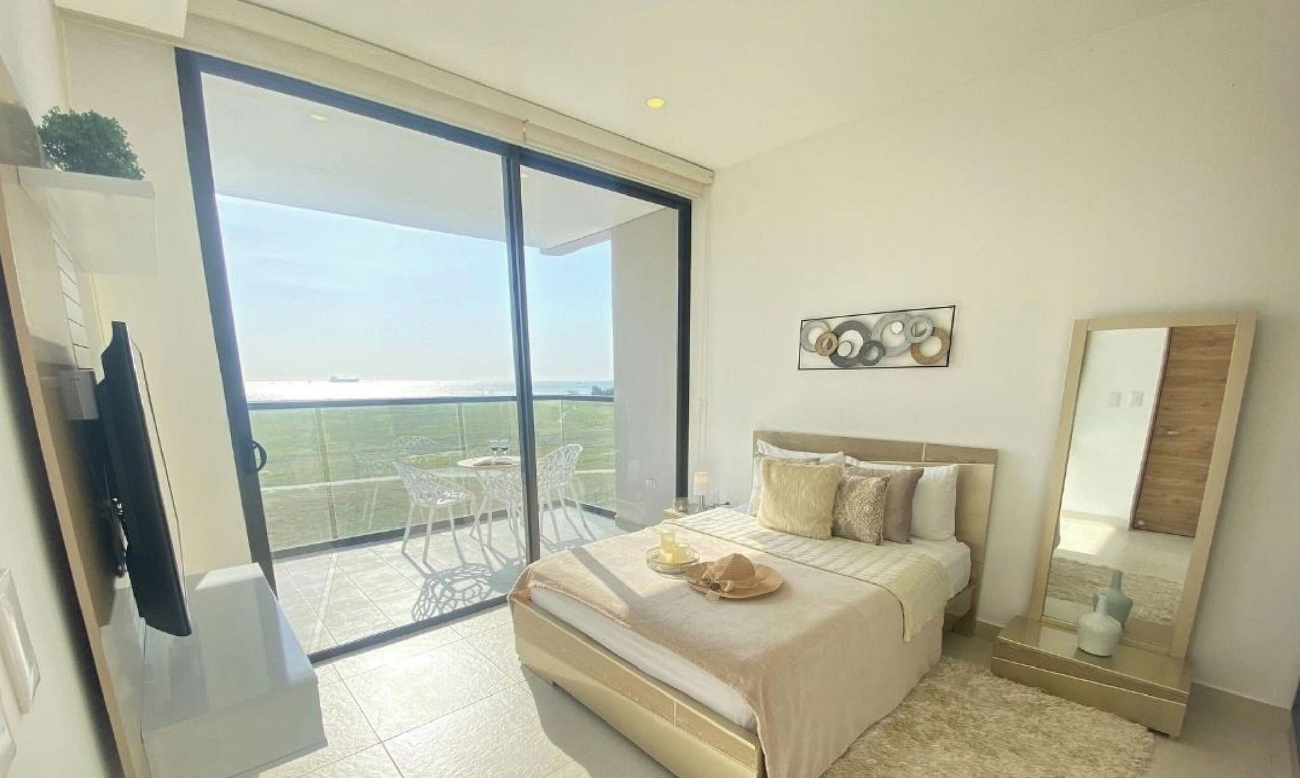 A bedroom with a bed, a bedside table, a mirror, and a view of the outdoors through a sliding glass door.