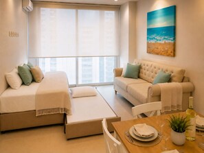 A hotel room with a bed, a sofa, a dining table with chairs, a window with blinds, and a painting on the wall.