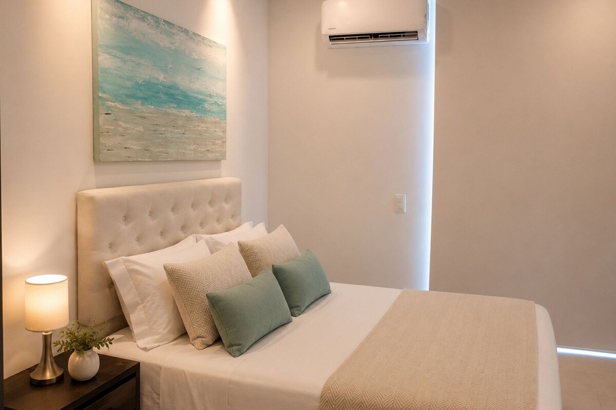A bedroom with a bed, a nightstand, a lamp, a wall-mounted air conditioner, and a painting.
