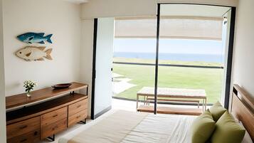 Une chambre à coucher avec un lit, une commode en bois, une petite table avec un bol, un vase de fleurs et une vue sur la plage par une grande fenêtre.