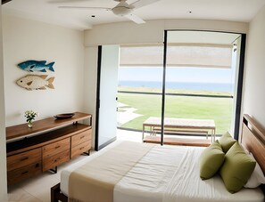 Un dormitorio con cama, cómoda de madera, una mesita con un frutero, un jarrón con flores y vista a la playa a través de un ventanal grande.