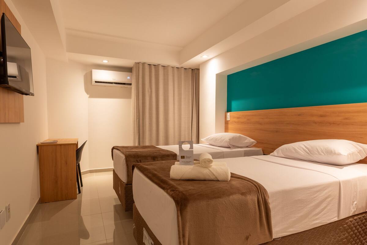 A hotel room with a bed, a desk, a chair, a TV, and an air conditioning unit.