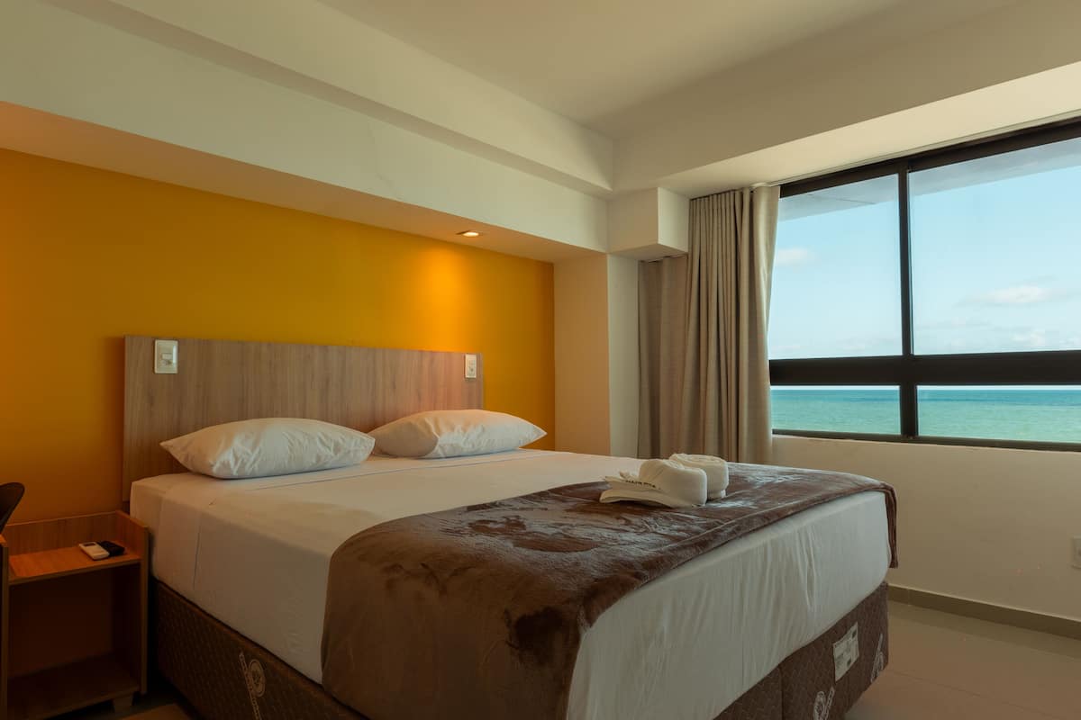 A hotel room with a bed, a desk, and a window with a view of the sea.