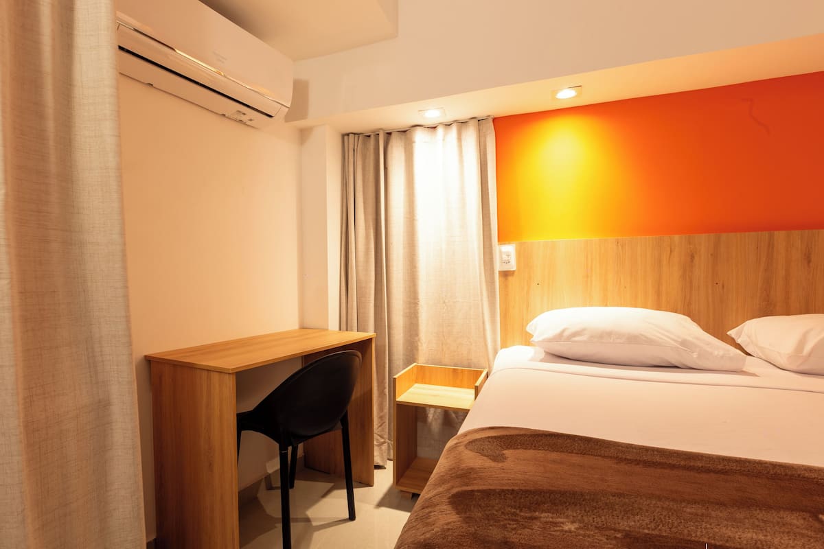 A hotel room with a wooden desk, a chair, a bed with white pillows, and an orange accent wall.