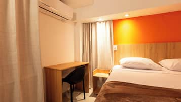Quarto de hotel com escrivaninha de madeira, uma cadeira, cama com almofadas brancas e uma parede laranja.