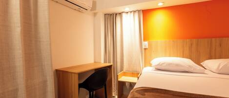 Quarto de hotel com escrivaninha de madeira, uma cadeira, cama com almofadas brancas e uma parede laranja.