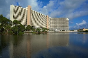 Un hotel grande con varios pisos, una masa de agua reflectante al frente y un cielo despejado.