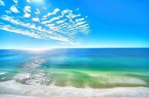 Uma praia com águas cristalinas, areal arenoso e céu azul com nuvens.