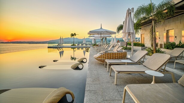 Une piscine à débordement paisible, dotée de chaises longues et de parasols, offrant une vue sur un coucher de soleil et des montagnes lointaines.