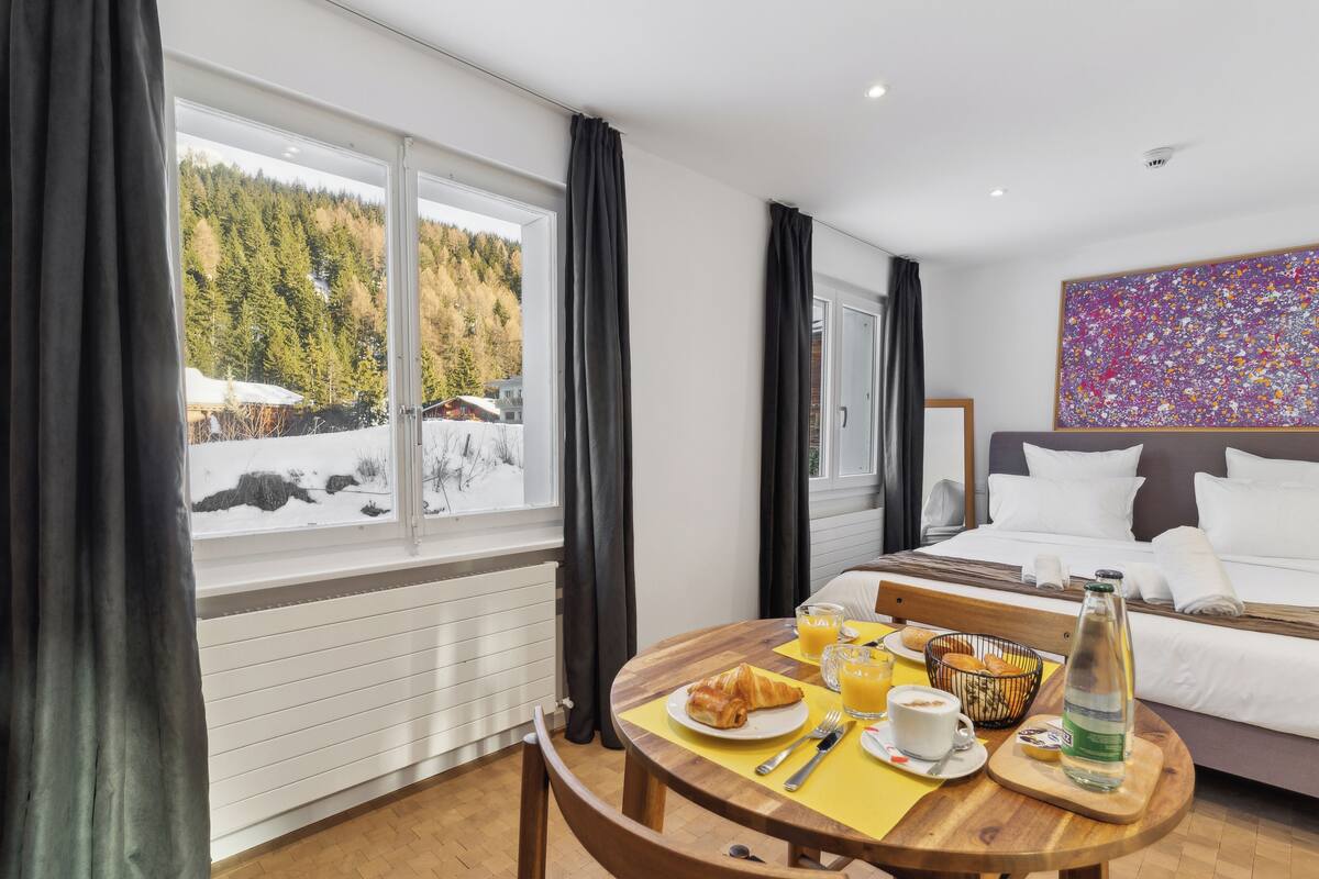 Una camera d'hotel con un letto, un tavolo con la colazione, una finestra vista alberi e neve, e un quadro astratto dai colori vivaci appeso al muro.