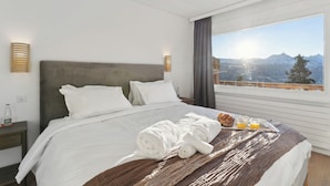 A neatly made bed with white linens, a brown throw blanket, and rolled towels. There is a breakfast tray with bread, juice, and a cup on the bed. The room has a large window with a mountain view and a view of the sun. - Crans Montana Suites (Crans-Montana)