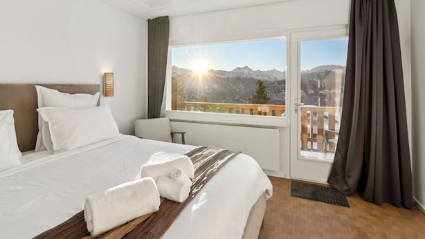 A hotel room with a bed, a nightstand, a window with a view of mountains, and a balcony. - Crans Montana Suites (Crans-Montana)