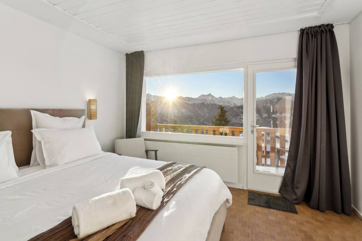 Una camera d'hotel con un letto, un comodino, una finestra vista montagne e un balcone.
