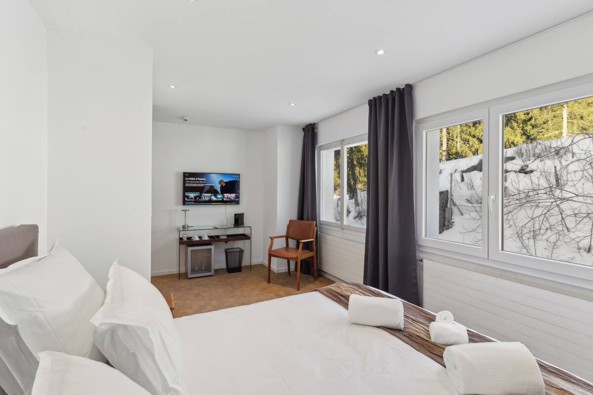 Una camera d'albergo con un letto, una scrivania con una sedia, una TV e una finestra vista alberi innevati.