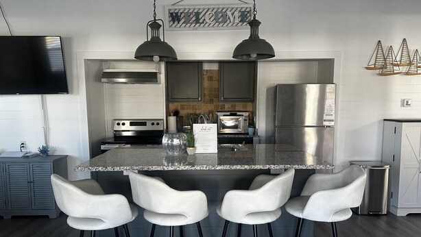 A modern kitchen with a central island, bar stools, stainless steel appliances, and a 'WELCOME' sign. - Ultimate Fishing Retreat: Spacious 3BR Bolivar Island Home with Dock & Cleaning Stations - Sleeps 12! (Bolivar Peninsula)