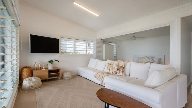 A modern living room with a white sofa, a flat-screen TV, a wooden side table, and a large window with blinds.
