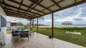 A covered patio with a view of the beach, outdoor furniture, and a table with a pitcher.