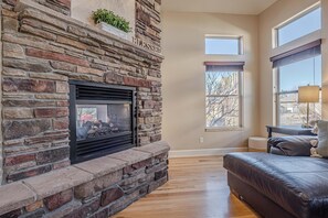 Ein Kamin aus Stein mit Feuer, ein Ledersofa und ein Fenster mit Blick auf Bäume.