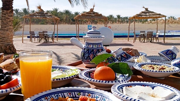 Una mesa de comedor con diversos platos, una tetera y un vaso de jugo de naranja, con el mar y palmeras de fondo y sombrillas.