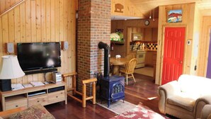 A cozy cabin interior with a TV, wooden furniture, a fireplace, and a kitchen area.