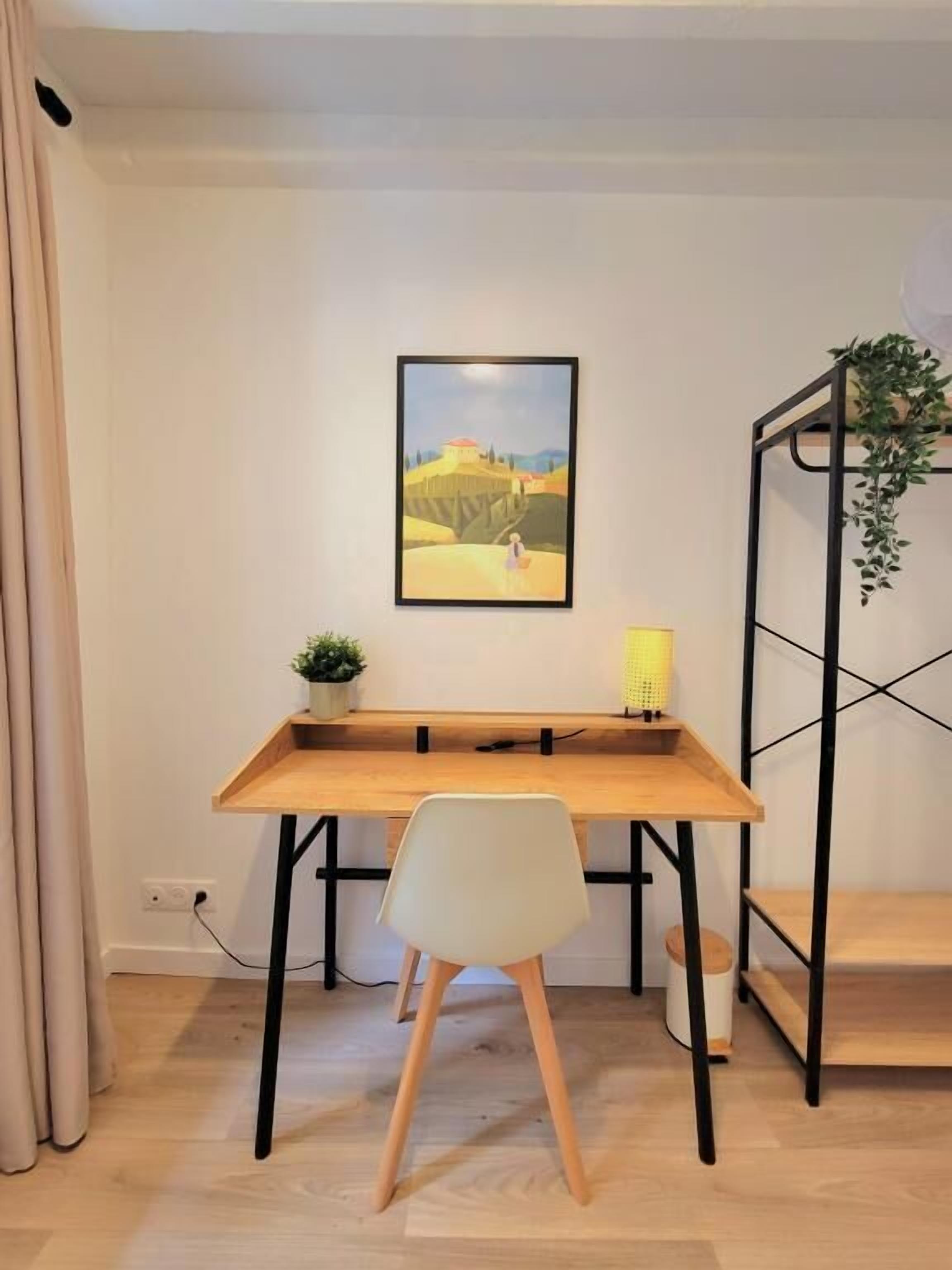 A minimalist workspace with a wooden desk, a white chair, a framed painting, a small plant, and a black metal shelf with a hanging plant.