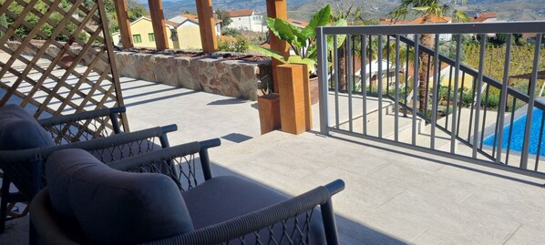 A balcony with wicker chairs, a stone planter, and a view of a swimming pool and distant houses. - Quinta de Raj (Peso da Régua)