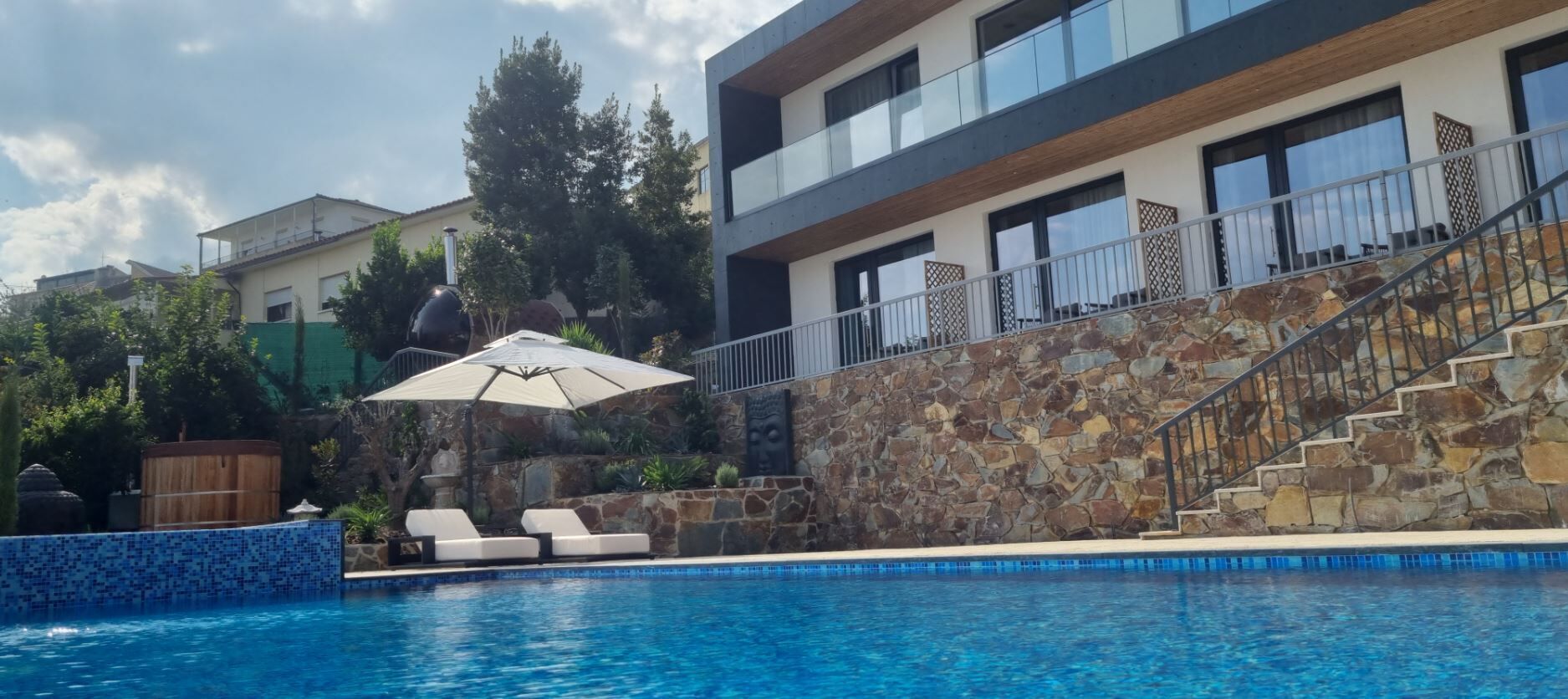 A modern pool area with a stone wall, a staircase, and a lounge chair under an umbrella.