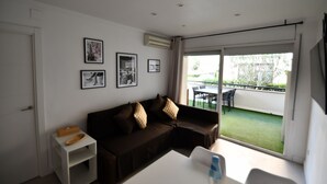 Un salón moderno con un sofá marrón, una mesita blanca y un ventanal de vidrio que da a un espacio exterior con una mesa y sillas.