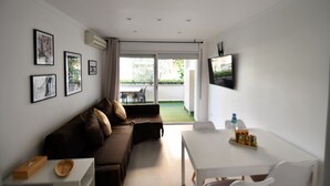 Una sala moderna con un sofá marrón, una mesa blanca y un televisor montado en la pared. Hay cuadros enmarcados en la pared, y una puerta corrediza de vidrio da a un balcón con una mesa y sillas afuera.