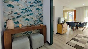 Un salon moderne avec un papier peint à motifs floraux, une console en bois, deux fauteuils confortables, un téléviseur posé sur un meuble en bois, et un coin repas avec des chaises et une table.