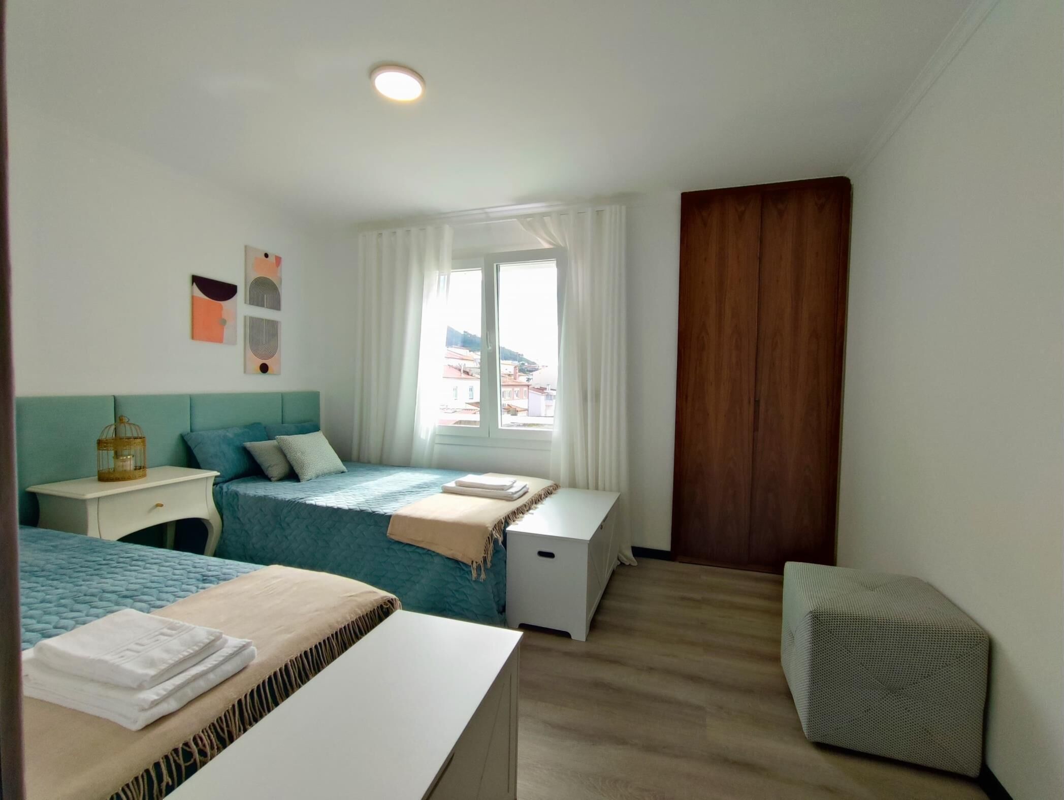 A hotel room with a bed, a bedside table, a small bedside cabinet, a window with curtains, and a view of buildings outside.