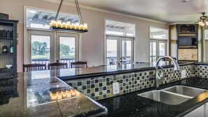 A modern kitchen with a large island, black countertops, and a tiled backsplash. There are two windows providing natural light and a view of the outdoors.