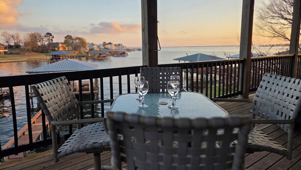 A wooden deck with a glass table and wicker chairs overlooking a body of water with boats and buildings in the background.