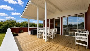 Una terraza techada al aire libre con suelo de madera, mobiliario blanco y un amplio ventanal corredera que da a un espacio interior.