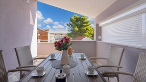 Un balcón con sillas de mimbre, una mesa para dos con tazas y un jarrón de flores, y vistas a edificios y árboles.