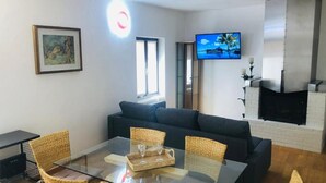 A modern living room with a glass dining table, wicker chairs, a dark sofa, a flat-screen TV mounted on the wall, and a fireplace. - LA TERRAZZA DI AIDA (Verona)