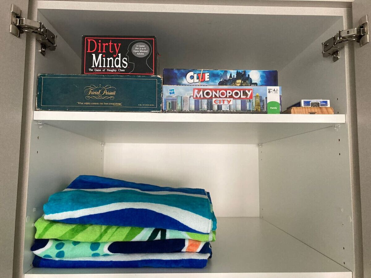 Een bergkast met twee planken. Op de bovenste plank staan bordspellen, waaronder 'Dirty Minds', 'Clue' en 'Monopoly City'. De onderste plank bevat opgevouwen handdoeken in verschillende kleuren.