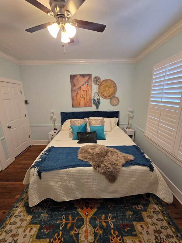 A bedroom with a bed, a ceiling fan, a painting, and a patterned rug.