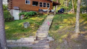 A log cabin with a porch, a driveway, and a small garden. - Charming 2-bedroom cottage lake property with kayaks firepit and Great fishing! (Elcho)