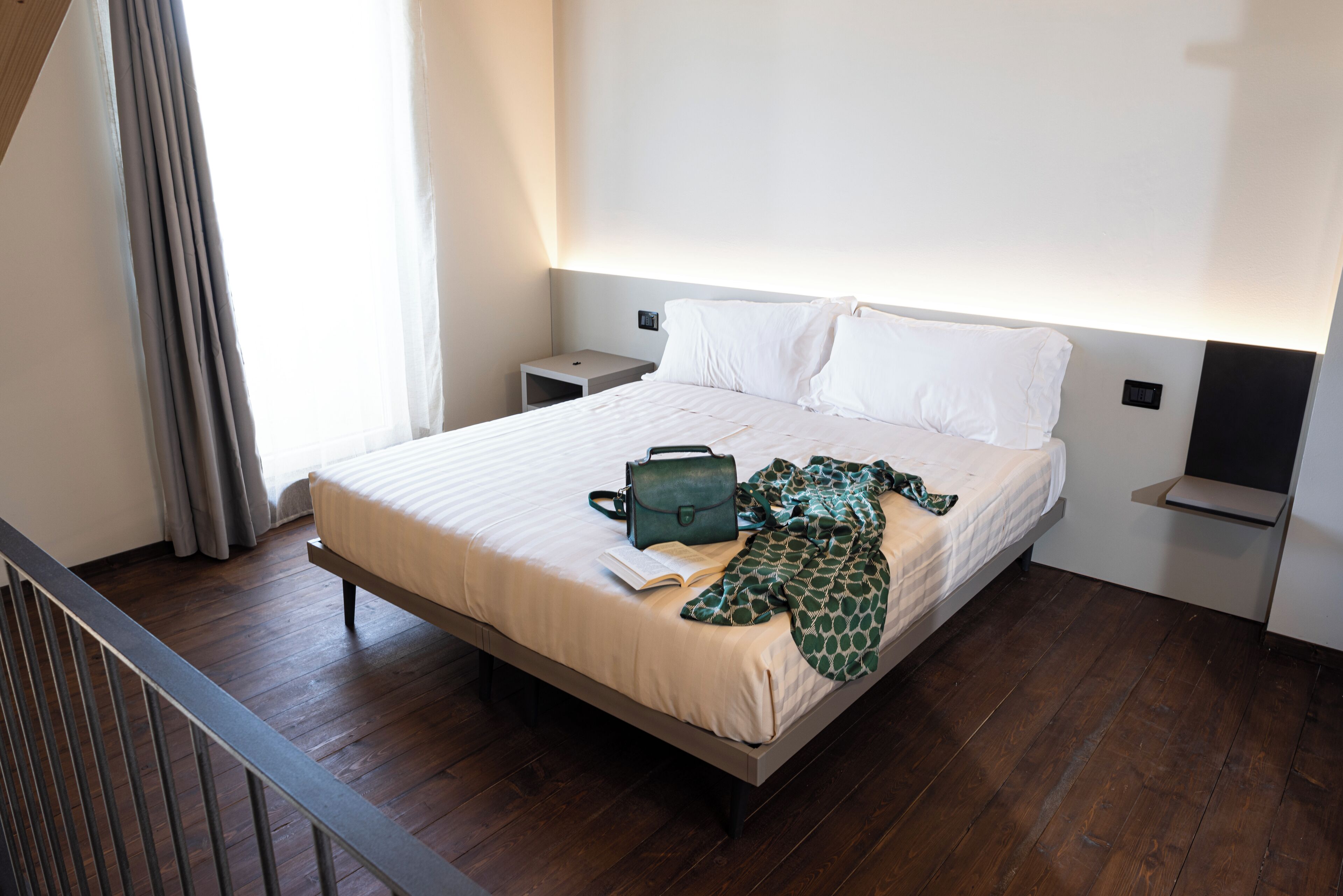A neatly made bed with a green bag and a book on it, a bedside table, and a nightstand.