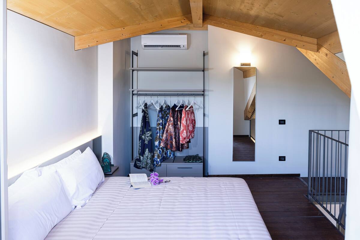 A bedroom with a bed, a closet with hanging clothes, a staircase, and an air conditioning unit.