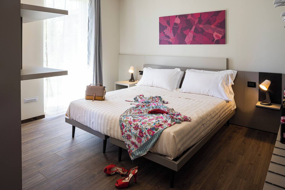 A modern hotel room with a large bed, bedside tables, a nightstand, and a window with curtains.