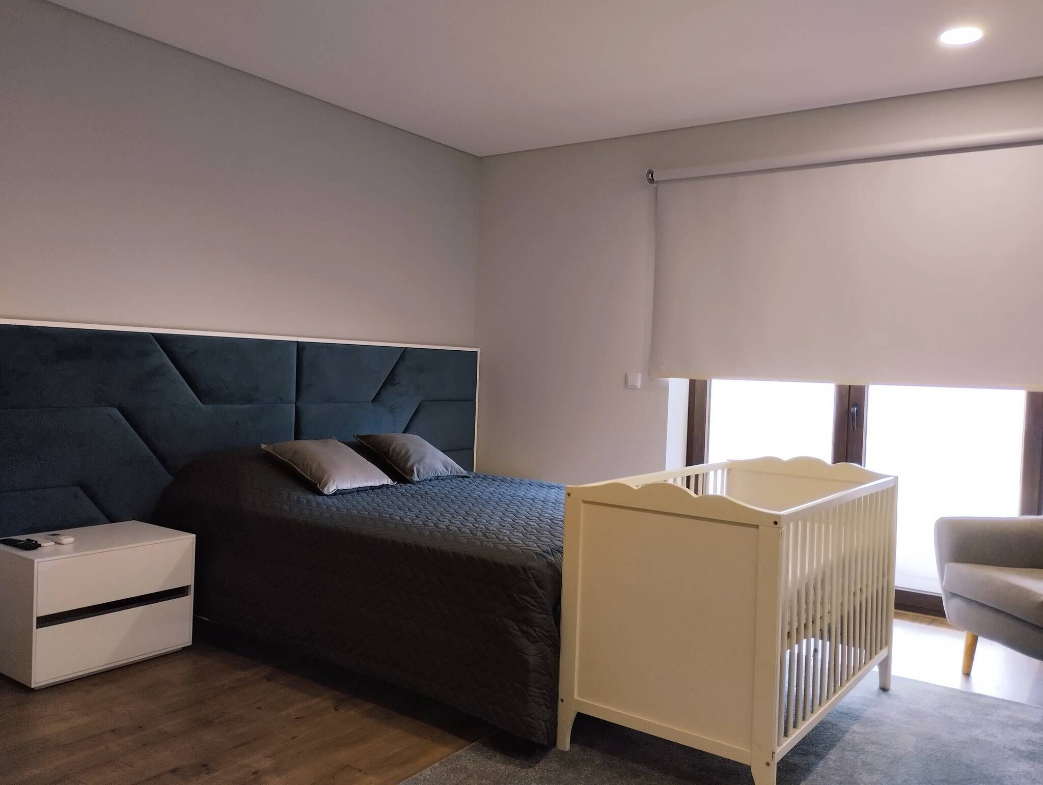 A modern bedroom with a large bed, a white nightstand, a white crib, and a window with a view.