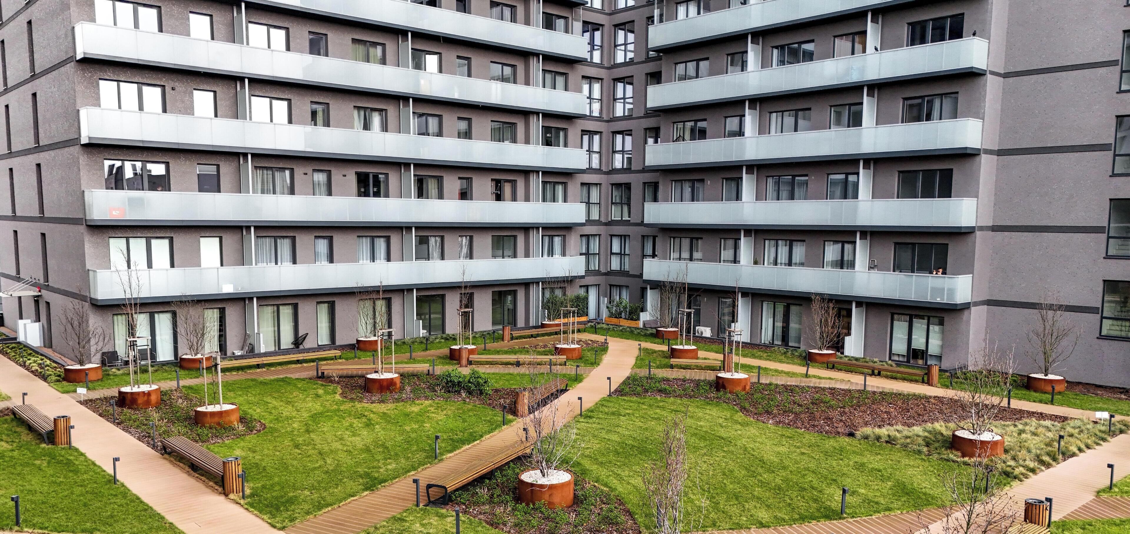 Ein modernes, mehrgeschossiges Gebäude mit einem Innenhof, der Wege, Bänke und Pflanzgefäße zeigt.